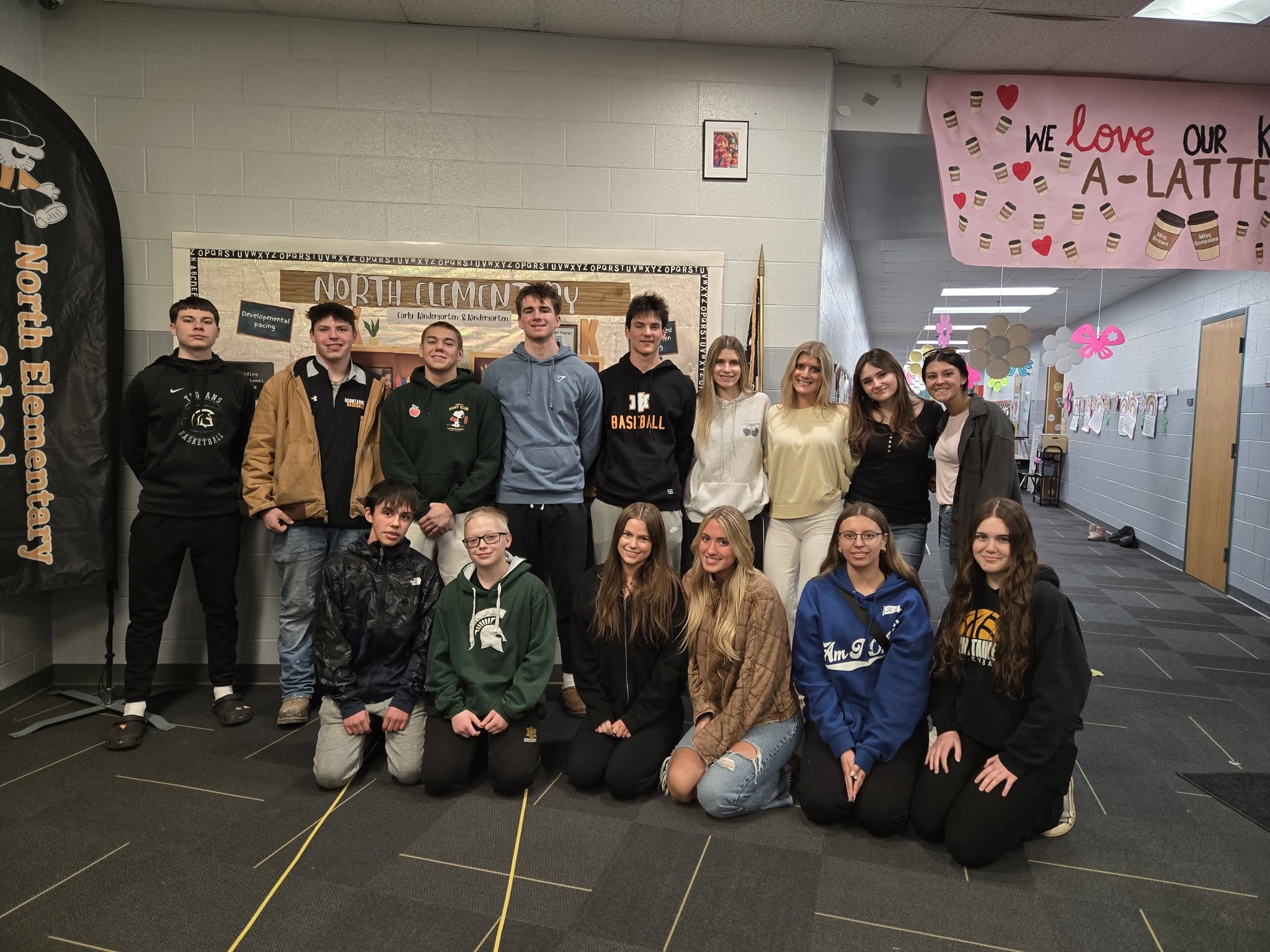 A group of 15 North Elementary and Iron Mountain High School students pose together in a school hallway in front of a bulletin board that reads "North Elementary." Six students kneel in the front row, while nine stand in the back row.